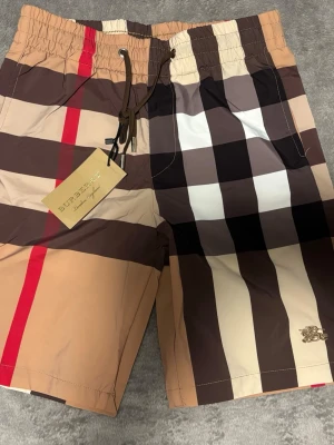 Rutig shorts från Burberry - Säljer ett par klassiska rutiga shorts från Burberry i beige, brunt, svart, vitt och rött. Shortsen har snörning i midjan och den ikoniska Burberry-loggan på benet. Perfekta för sommaren och har en loose passform.