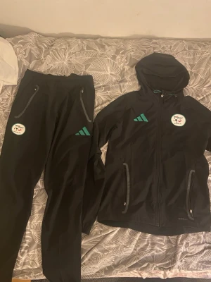 Svart Adidas träningsset med luva - Säljer ett svart träningsset från Adidas med jacka och byxor. Jackan har huva, dragkedja och två fickor med dragkedja. Både jacka och byxor har gröna Adidas-loggor och ett runt klubbmärke på bröst och lår. Materialet är lätt och funkar perfekt till träning.