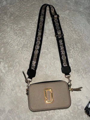 Beige axelväska Marc Jacobs - Snygg beige axelväska från Marc Jacobs med justerbar svart axelrem. Väskan har guldiga metalldetaljer, dubbla dragkedjor och är tillverkad i strukturerat läder. Perfekt för att bära det viktigaste med stil.