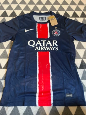 PSG matchtröja Nike marinblå röd - Snygg PSG matchtröja från Nike i marinblått med en bred röd och vit rand mitt på. Tröjan har klubbmärke och Nike-logga på bröstet samt Qatar Airways-tryck. Perfekt för fotbollsfans som vill visa sitt stöd för Paris Saint-Germain.