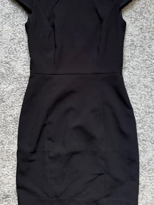 Svart kortärmad pennklänning - Stilren svart pennklänning med korta ärmar och figurnära passform. Klänningen har en klassisk siluett och dragkedja i ryggen. Perfekt för dig som gillar enkel och tidlös design.