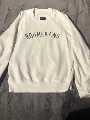 Vit tröja  - Snygg vit sweatshirt från Boomerang med svart tryck över bröstet. Tröjan har rund halsringning, ribbade muddar och är tillverkad i mjukt bomullsmaterial. Perfekt för en avslappnad och stilren look. Den har två små hål men hålen sitter inåt mot tröjan så det är inget som syns. 