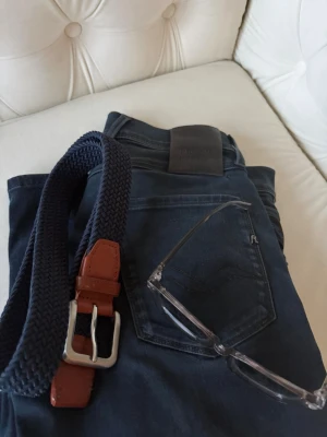 Flätat bälte  - Snyggt mörkblått flätat bälte med bruna läderdetaljer och silverfärgat spänne. Perfekt accessoar för att lyfta din outfit med en klassisk och stilren touch. Bältet är flexibelt och passar till både jeans och chinos.
