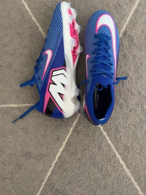 Nike Mercurial blå fotbollsskor - Säljer ett par Nike Mercurial fotbollsskor i blått med vita och rosa detaljer samt svart grafik på sidan. Skorna har snörning, syntetiskt material och dobbar för bra grepp på planen. Perfekta för dig som vill sticka ut på fotbollsplanen.