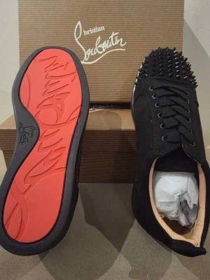 Svarta sneakers med nitar Louboutin - Säljer ett par svarta sneakers från Christian Louboutin med mattsvarta nitar på tån och den ikoniska röda sulan. Skorna har snörning och är tillverkade i mocka för en lyxig känsla. Perfekta för dig som vill sticka ut med en edgy stil.