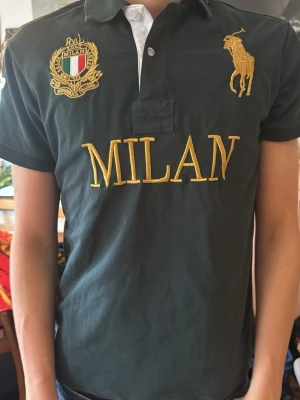 Mörkgrön Milan t-shirt från Ralph Lauren - Snygg grön t-shirt från Ralph Lauren med stora guldfärgade broderier, MILAN-text på bröstet och klubbmärke. Klassisk pikékrage och knappslå framtill, samt vit siffra 5 på ärmen. Perfekt för dig som gillar sportig och stilren look. Den är köpt på loppis så äkthet kan ej garanteras