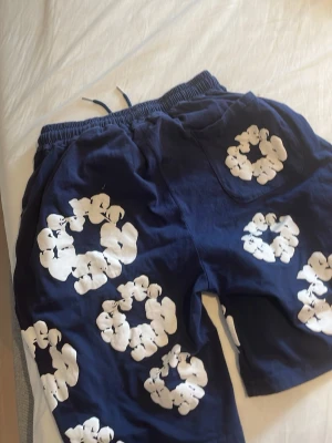 Blå shorts med vita blommor - Säljer ett par blå shorts med stora vita blommönster. De har resår i midjan med snörning och är tillverkade i ett mjukt bomullstyg. Perfekta för sommardagar och har en avslappnad passform.