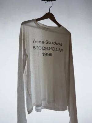 Vit långärmad topp Acne Studios - Vit långärmad topp från Acne Studios med tryck 'Acne Studios STOCKHOLM 1996' på bröstet. Tunn och lätt i materialet, perfekt för en clean och enkel stil. Rund halsringning och relaxed fit.