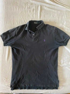 Svart pikétröja från Polo Ralph Lauren - Klassisk svart pikétröja från Polo Ralph Lauren med lila broderad logga på bröstet. Tröjan har korta ärmar, krage och knappar vid halsen. Perfekt till både jeans och shorts för en clean och stilren look.