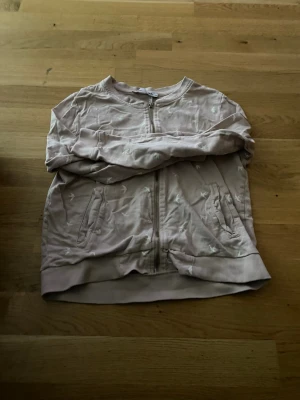 Ljusrosa zip hoodie från 157 - Ljusrosa hoodie med dragkedja från 157. Tröjan har vita fågelmönster, två fickor framtill och ribbade muddar vid ärmslut och nederkant. Tillverkad i mjukt bomullsmaterial och har en avslappnad passform som passar perfekt till vardagslooken.