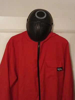 Squid Game maskeradoutfit röd - Röd helkroppsdräkt med huva och svart dragkedja framtill, inspirerad av Squid Game. Tillhörande svart mask med vitt cirkelmönster. Perfekt för cosplay eller maskerad. Dräkten har en bröstficka med Squid Game-logga.