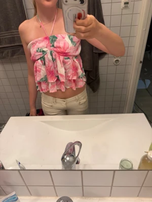 Blommig volang bandeau topp - Supersöt bandeau topp med rosa och vita blommor, volanger och halterneckband. Toppen är croppad och har en luftig passform, perfekt för varma dagar. Materialet känns lätt och somrigt.