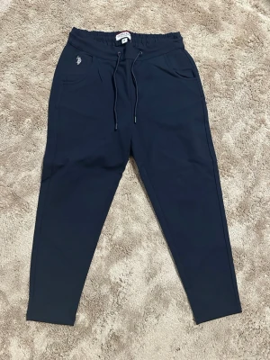 Mörkblå mjukisbyxor från U.S POLO ASSN - Snygga mörkblå mjukisbyxor från Polo med dragsko i midjan och två fickor fram. Byxorna har en liten broderad logga på vänster sida och är tillverkade i mjukt, bekvämt material. Perfekta för chill dagar eller när du vill vara extra bekväm. Storlek M