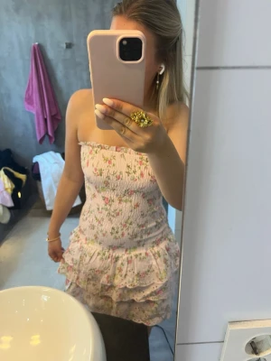 Rosa blommig kort klänning med volang - Söt axelbandslös kort klänning i ljusrosa med blommigt mönster. Helt ny med prislappen kvar. Säljer för att jag köpt en annan klänning.