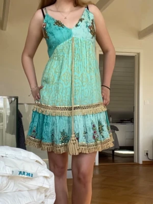 Turkos kortklänning med volang - Säljer en turkos kortklänning i boho-stil med broderade blommor, volangkant och dekorativa tofsar framtill. Klänningen har smala axelband, v-ringning och detaljer i beige. Perfekt för sommarens alla tillfällen! Köpt på marknad i Spanien för 30€, aldrig använd med lapp kvar💕