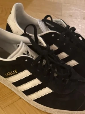 Svarta Adidas Gazelle sneakers - Adidas Gazelle sneakers i svart mocka med vita detaljer och klassiska tre ränder på sidan.  Storlek 38,5. Använda men i bra skick