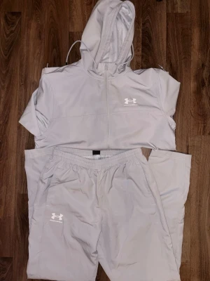 Grått träningsset från Under Armour - Finns kvitto på den.!!! Snyggt grått träningsset från Under Armour med vattenavvisande material. Setet består av en vindjacka med huva och dragkedja samt matchande byxor med resår i midjan. Perfekt för träning utomhus eller chill dagar.