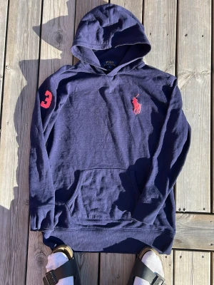 Marinblå hoodie från Polo Ralph Lauren - Snygg marinblå hoodie från Polo Ralph Lauren med röd logga på bröstet och en röd siffra 3 på ärmen. Klassisk känguruficka framtill och huva. Perfekt för en avslappnad och sportig stil.OBS barnstorlek 