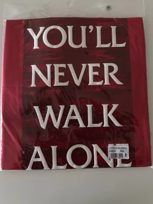 Liverpool röd t-shirt YNWA - Röd t-shirt från Liverpool FC med det ikoniska trycket 'You'll Never Walk Alone' i vitt på framsidan. Tillverkad i mjuk bomull och perfekt för alla fans som vill visa sitt stöd för laget. Klassisk passform och officiell supporterprodukt.