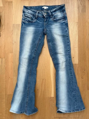 Blå bootcut jeans med slitningar - Säljer ett par blå bootcut jeans med snygga slitningar och ljusa tvättade partier. Jeansen har klassisk femficksmodell, låg midja och utsvängda ben. Perfekta för en avslappnad och trendig look.