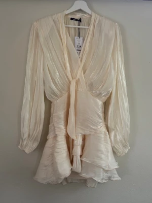 Krämfärgad omlottklänning Gina Tricot - Säljer en krämfärgad kort omlottklänning från Gina Tricot med långa ärmar och v-ringning. Klänningen har volangdetaljer och ett knytband i midjan. Materialet är glansigt och lätt, vilket ger ett elegant intryck.