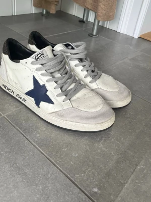 Golden Goose ball stars - Snygga vita sneakers från Golden Goose med en mörkblå stjärna på sidan och grå skosnören. Skorna har svarta detaljer vid hälen och textdetaljer på sidan. Ovandelen är i läder och mocka, med klassisk låg modell och slitna detaljer för en cool look.