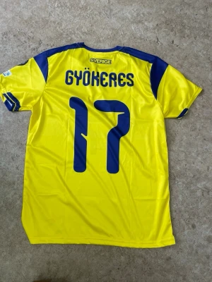 Sverige matchtröja Gyökeres #17 - Gul och blå officiell matchtröja från svenska landslaget med Gyökeres och nummer 17 på ryggen. Tröjan har korta ärmar, blå detaljer på axlar och ärmslut samt broderad landslagslogga framtill. Tillverkad i lätt och ventilerande polyester, perfekt för fotboll.