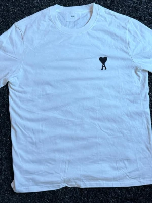 Vit t-shirt från Ami med hjärtdetalj - Vit t-shirt från Ami med svart hjärta och bokstaven A tryckt på bröstet. Klassisk rund halsringning och korta ärmar. Tillverkad i mjuk bomull för en skön känsla och enkel stil.