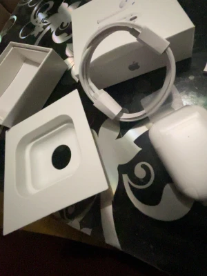Apple AirPods med laddningsetui - Säljer ett par vita Apple AirPods med tillhörande laddningsetui och originalförpackning. Hörlurarna är trådlösa och har en stilren, minimalistisk design i plast. Perfekta för musik, samtal och vardagsbruk. Laddningskabel och box ingår.