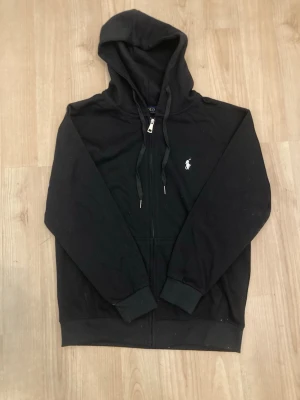 Svart hoodie från Polo Ralph Lauren - Klassisk svart hoodie från Polo Ralph Lauren med dragkedja framtill och vit broderad logga på bröstet. Hoodien har justerbar huva med snören och ribbade muddar vid ärmar och nederkant. Tillverkad i mjukt bomullsmaterial för en skön känsla. Storlek L