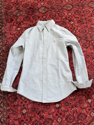 Ljusgrå skjorta från Ralph Lauren - Klassisk ljusgrå skjorta från Ralph Lauren med button-down krage och broderad logga på bröstet. Skjortan har lång ärm, normal passform och är tillverkad i mjuk bomull. Perfekt till både jeans och chinos för en clean look.