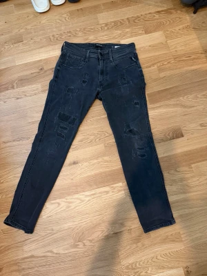 Svarta slitna jeans från Replay - Säljer ett par svarta skinny jeans från Replay med slitningar och hål på benen. Jeansen har fem fickor, bälteshällor och klassisk knappgylf. Snygg tvättad look och smal passform som sitter tajt längs benen.