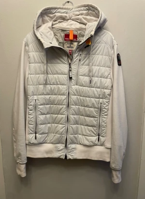 Vit pufferjacka från Parajumpers - Snygg vit pufferjacka från Parajumpers med quiltad front och huva. Jackan har dragkedja framtill, två sidofickor med dragkedja och en bröstficka. Ärmar och rygg är i mjuk fleece, vilket ger en sportig och modern look. Parajumpers-logga på ärmen.
