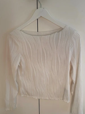 Vit långärmad topp med struktur - Snygg vit långärmad topp med vågigt strukturerat mönster. Tunn och lätt, perfekt att styla med olika outfits. Toppen har en rund halsringning och är figurnära i modellen.