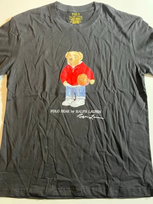 Svart Polo Bear t-shirt från Ralph Lauren - Cool svart t-shirt från Ralph Lauren med Polo Bear-tryck framtill. Björnen har på sig en röd hoodie, blå jeans och håller en basketboll. Klassisk rund hals och korta ärmar. Perfekt för dig som gillar streetstyle och ikoniska prints.