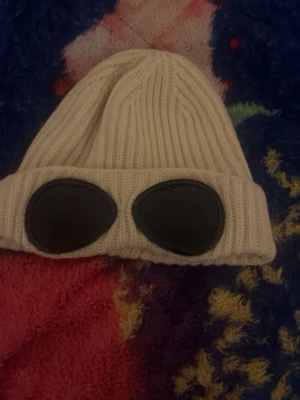 Vit ribbstickad mössa med goggles - Säljer en vit ribbstickad mössa med uppvikt kant och inbyggda svarta goggles framtill. Mössan har en cool streetwear-vibe och är perfekt för dig som vill sticka ut. Materialet känns mjukt och stretchigt, vilket gör den bekväm att bära hela dagen. CP company mössa.