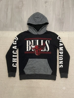 Chicago Bulls hoodie NBA svart/grå - Snygg hoodie i svart och grått med Chicago Bulls-logga och texten 'CHICAGO' och 'CHAMPIONS' på ärmarna. Stor ficka framtill och kontrasterande huva. Perfekt för dig som gillar basket och vill ha en sportig stil.