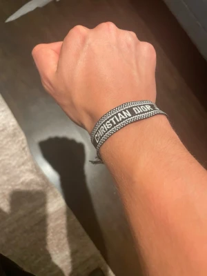 Christian Dior svart tygarmband - Snyggt svart tygarmband från Christian Dior med vit broderad text och dekorativa fransar i ändarna. Armbandet har en justerbar knytning och en cool, trendig look som passar perfekt till streetwear eller för att lyfta din outfit.