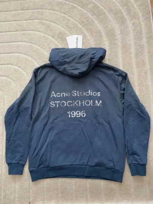 Blå hoodie från Acne Studios - Snygg blå hoodie från Acne Studios med tryck på ryggen där det står 'Acne Studios STOCKHOLM 1996'. Hoodien har huva, långa ärmar och ribbade muddar. Tillverkad i mjukt bomullsmaterial och har en clean look utan fickor framtill.
