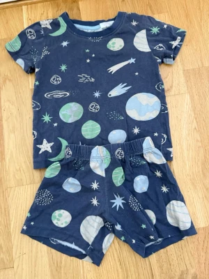 Blått rymdset från H&M, stl 92 - Säljer ett blått tvådelat set från H&M i storlek 92. Setet består av en t-shirt och shorts med coolt rymdtryck i grönt, vitt och blått med planeter, stjärnor och kometer. Tillverkat i mjuk bomull, perfekt för chill dagar hemma eller som pyjamas.