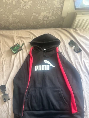Svart hoodie med röd detalj och tryck - Säljer en svart hoodie med röda detaljer längs ärmarna och ett stort vitt tryck framtill som liknar PUMA-loggan fast med texten 'PMMN'. Hoodien har huva med snören och en stor magficka. Perfekt för dig som gillar streetwear och vill sticka ut. Pris kan diskuteras.