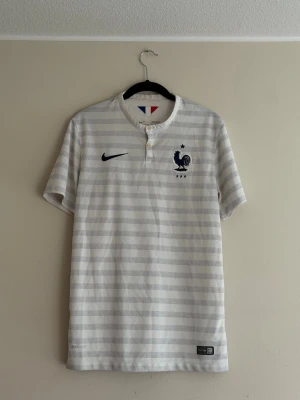 Frankrike fotbollströja Nike VM 2014 - Säljer en vit och ljusgrå randig fotbollströja från Nike med Frankrikes landslagsemblem på bröstet. Tröjan har korta ärmar, två knappar vid halsen och är tillverkad i ett lätt, andningsbart material perfekt för träning eller match.