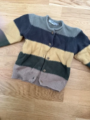 Randig stickad kofta från mywear - Stickad kofta med breda ränder i grått, mörkgrönt, beige och gul. Tröjan har rund halsringning, träknappar framtill och grovstickad struktur. Perfekt för lager på lager och ger en chill vibe till outfiten.
