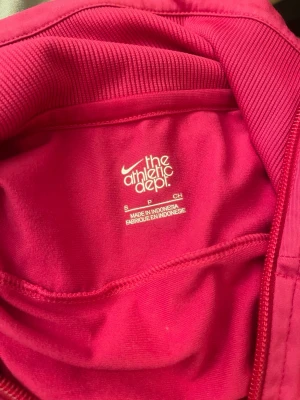Rosa ziptröja från Nike - Snygg rosa ziptröja från Nike med vit och svart detalj över bröstet. Tröjan har hög krage, ribbade muddar och två fickor framtill. 