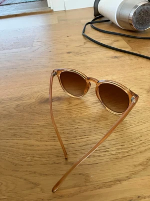 Bruna runda solglasögon i plast - Snygga solglasögon med rundad form och transparent brun plastbåge. Glasen är mörkbruna och ger en cool vintagekänsla. Perfekta för att lyfta din stil under soliga dagar.
