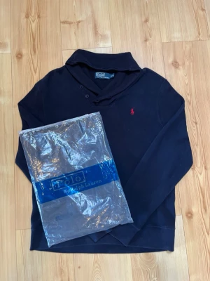 Mörkblå hoodie från Polo Ralph Lauren - Snygg mörkblå hoodie från Polo Ralph Lauren med klassisk röd logga broderad på bröstet. Hoodien har en huva och knappar vid halsen för extra stil. Tillverkad i mjukt material som känns skönt mot huden. Perfekt för en avslappnad och trendig look.