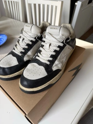 Svarta och vita MOA Concept sneakers - Snygga sneakers från MOA Concept i svart och vitt läder med kontrasterande detaljer. Skorna har vita snören, perforerad tå och en broderad W-logga på sidan. Klassisk siluett med retrovibbar och robust sula. Perfekta för dig som gillar streetwear och stilrena detaljer.