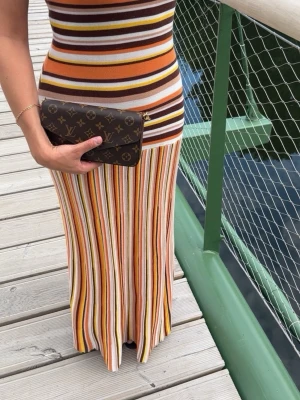 Randig långklänning med smala axelband - Säljer en långklänning med smala axelband och färgglada ränder i brunt, orange, gult, vitt och beige. Klänningen är figurnära och har en mjuk, stretchig känsla. Perfekt för sommarens alla tillfällen!