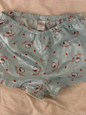 Hello Kitty pyjamasshorts i satin - Supersöta ljusblå pyjamasshorts med Hello Kitty-tryck och små röda hjärtan över hela. Shortsen har resår i midjan och är gjorda i ett glansigt satinliknande material som känns mjukt mot huden. Perfekta för mysiga hemmakvällar eller sommarens varma nätter.