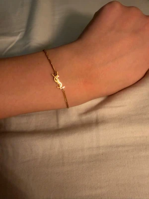 Saint Laurent armband  - Snyggt och tunt armband i guldfärgad metall med den ikoniska YSL-logotypen som detalj. Armbandet har en tunn kedja och justerbar längd med ett litet lås. Perfekt accessoar för dig som gillar stilrena och lyxiga smycken.